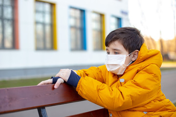 Portrait of a sad boy in a yellow jacket in a white medical mask sitting on a bench on the street