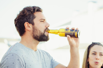 Wall Mural - Focused bearded guy enjoying beer with friends on outdoor terrace. Young man and woman in casual meeting outside. Beer party on rooftop concept