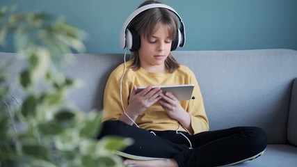 Wall Mural - Video of little girl listening to music with headphones and digital tablet while sitting on sofa at home.