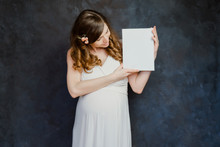 Pregnant Young Woman In White Dress Looking At Blank Canvas In Her Hands. Girl With Mockup Poster Frame On Dark Backdrop.