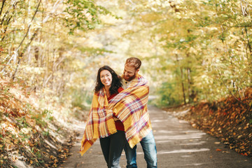 Beautiful couple man woman in love. Boyfriend and girlfriend wrapped in yellow blanket plaid hugging in a park on autumn fall day. Tenderness and happiness. Authentic real people.