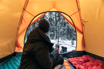 Wall Mural - man traveler wearing winter coat with sitting and holding a coffee cup in orange tent on campsite at