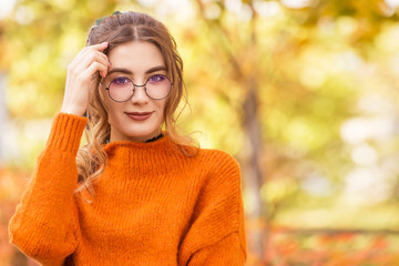 Beautiful young woman on a street in a park in October. The girl is dressed in sweater and in glass, round glasses. Student teacher is relaxing, walking in the park.