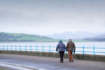 Senior couple walking at sea esplanade during rain storm in cold months