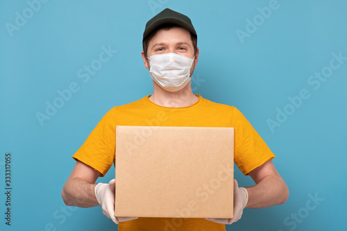 Fotografía Young delivery man in nedical mask holding and carrying a cardbox on blue wall