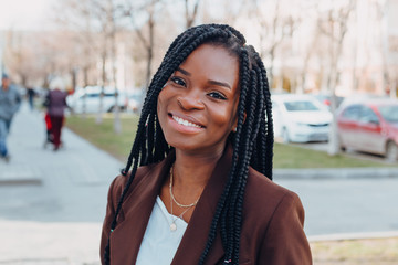 Wall Mural - Close up portrait of a beautiful young african american woman with pigtails hairstyle in a brown business suit walks along spring streets