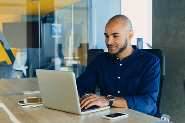 handsome cheerful african american man in creative atmosphere using laptop sitting on a wooden table