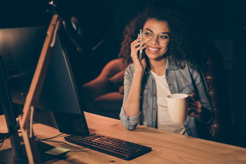 Poster - Photo of pretty young dark skin business lady look big screen monitor working overtime drink hot coffee talk telephone partner wear specs casual shirt sit table chair night office indoors