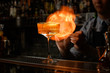 © fesenko - Glass of alcoholic drink and ice stands on bar counter. Bartender makes fire flame behind it.