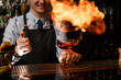 © fesenko - young smiling bartender holds glass with cocktail, spraying on it and sets on fire.