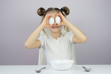 Portrait Of A Little Girl Holding Two Chicken Eggs Near Her Eyes And Smiling. Eggs Instead Of Eyes