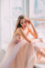 Portrait Beautiful Mysterious Girl With Long Blond Hair In Luxurious Airy Elegant Pastel Beige Dress Natural Cosmetics. Princess Sits On Windowsill, Hand Touches Head Backdrop White Window. Prom Party