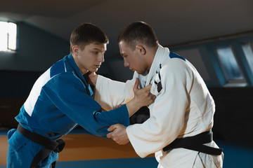 Two young judo caucasian fighters in white and blue kimono with black belts training martial arts in the gym with expression, in action, motion. Practicing fighting skills. Overcoming, reaching target