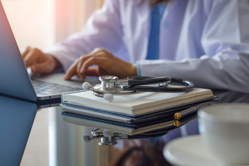 Female doctor in white lab coat typing on laptop computer with notebook and medical stethoscope on the desk at workplace. Medical technology ,Electronic health record system (EMR) concept