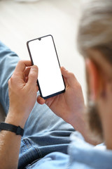 Millennial man user holding smart phone mock up white screen in hand using mobile dating app, check social media, order food delivery, reading news sit on couch at home. Over shoulder vertical view.