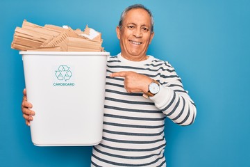 Wall Mural - Senior man recycling holding trash can with cardboard to recycle over blue background very happy pointing with hand and finger