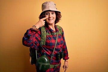 Poster - Middle age curly hair hiker woman hiking wearing backpack and water canteen using binoculars Pointing with hand finger to face and nose, smiling cheerful. Beauty concept