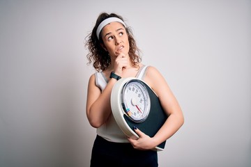 Sticker - Young beautiful woman with curly hair holding weighing machine over white background Thinking worried about a question, concerned and nervous with hand on chin