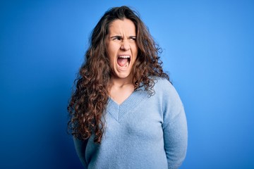 Sticker - Young beautiful woman with curly hair wearing blue casual sweater over isolated background angry and mad screaming frustrated and furious, shouting with anger. Rage and aggressive concept.