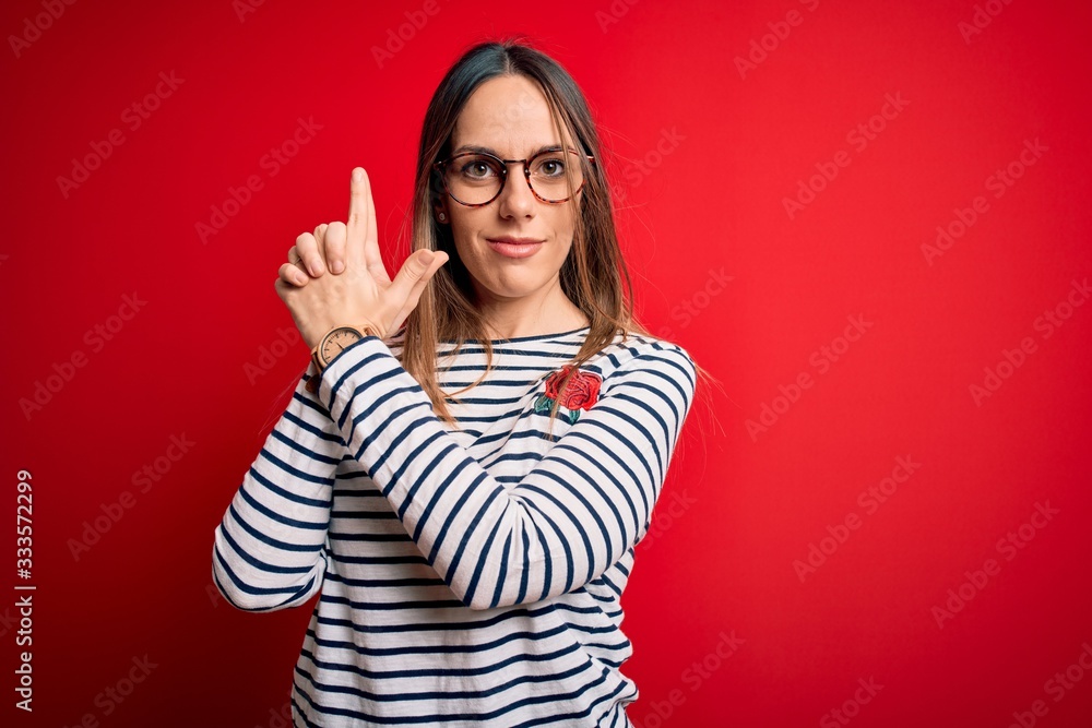 Young beautiful blonde woman with blue eyes wearing glasses standing over red background Holding symbolic gun with hand gesture, playing killing shooting weapons, angry face