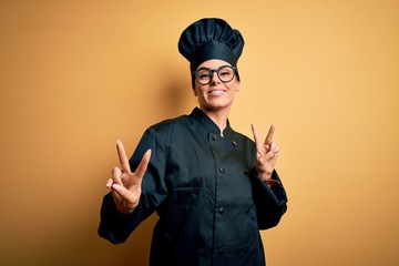 Sticker - Young beautiful brunette chef woman wearing cooker uniform and hat over yellow background smiling looking to the camera showing fingers doing victory sign. Number two.