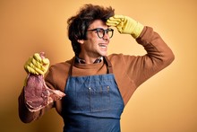 Young Handsome Butcher Man Holding Meet Steak Standing Over Isolated Yellow Background Very Happy And Smiling Looking Far Away With Hand Over Head. Searching Concept.