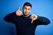 Young handsome man wearing casual sweater standing over isolated blue background Doing thumbs up and down, disagreement and agreement expression. Crazy conflict