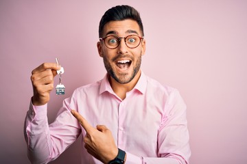 Young real estate business man holding new house keys over pink background very happy pointing with hand and finger