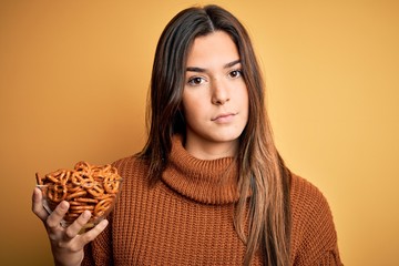 Poster - Young beautiful girl eating baked german pretzel standing over isolated yellow background with a confident expression on smart face thinking serious