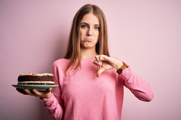 Wall Mural - Young beautiful redhead woman holding birthday cake over isolated pink background with angry face, negative sign showing dislike with thumbs down, rejection concept