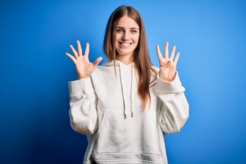 Young beautiful redhead sporty woman wearing sweatshirt over isolated blue background showing and pointing up with fingers number nine while smiling confident and happy.