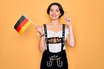 Wall Mural - Young patriotic german woman with blue eyes wearing octoberfest dress holding germany flag surprised with an idea or question pointing finger with happy face, number one