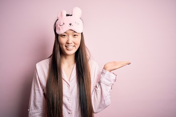 Sticker - Young asian woman wearing pajama and sleep mask over pink isolated background smiling cheerful presenting and pointing with palm of hand looking at the camera.