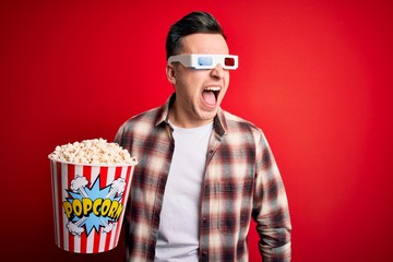 Wall Mural - Young handsome caucasian man wearing 3d movie glasses and eating popcorn angry and mad screaming frustrated and furious, shouting with anger. Rage and aggressive concept.