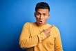 © Krakenimages.com - Young handsome latin man wearing yellow casual sweater over isolated blue background Surprised pointing with finger to the side, open mouth amazed expression.