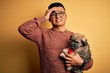 © Krakenimages.com - Young handsome latin man holding cute puppy pet over isolated yellow background stressed with hand on head, shocked with shame and surprise face, angry and frustrated. Fear and upset for mistake.