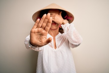 Canvas Print - Middle age brunette woman wearing asian traditional conical hat over white background covering eyes with hands and doing stop gesture with sad and fear expression. Embarrassed and negative concept.