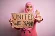 © Krakenimages.com - Young activist woman wearing pink muslim hijab holding banner with united we stand message with open hand doing stop sign with serious and confident expression, defense gesture