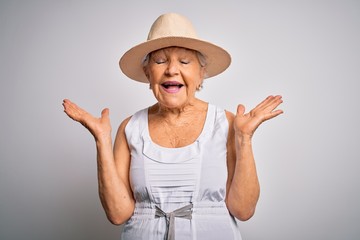 Senior beautiful grey-haired woman on vacation wearing casual summer dress and hat celebrating mad and crazy for success with arms raised and closed eyes screaming excited. Winner concept