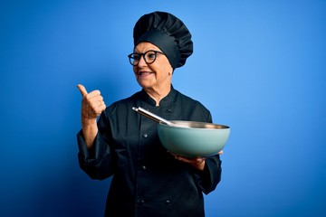 Wall Mural - Senior beautiful grey-haired chef woman wearing cooker uniform and hat using whisk and bowl pointing and showing with thumb up to the side with happy face smiling