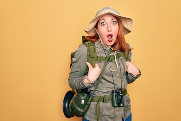 Poster - Young redhead backpacker woman hiking wearing backpack and hat over yellow background afraid and shocked with surprise and amazed expression, fear and excited face.