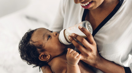 Portrait of enjoy happy love family african american mother playing with adorable little african american baby.Mom feeding bottle of milk to baby cute son in a white  bedroom.Love of black family 