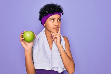 Beautiful african american afro sporty woman doing exercise wearing towel eating green apple serious face thinking about question, very confused idea