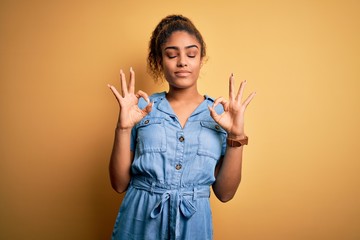 Wall Mural - Young beautiful african american girl wearing denim dress standing over yellow background relaxed and smiling with eyes closed doing meditation gesture with fingers. Yoga concept.