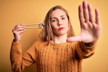 Sticker - Young beautiful blonde woman eating sushi using chopsticks over isolated yellow background with open hand doing stop sign with serious and confident expression, defense gesture