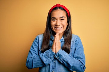 Poster - Young beautiful asian woman wearing casual denim shirt and diadem over yellow background praying with hands together asking for forgiveness smiling confident.