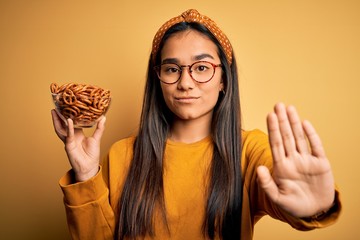 Wall Mural - Young beautiful asian woman holding bowl with german baked pretzels over yellow background with open hand doing stop sign with serious and confident expression, defense gesture
