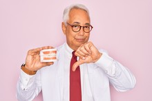 Middle Age Senior Grey-haired Dentist Man Holding Prosthesis Denture Over Pink Background With Angry Face, Negative Sign Showing Dislike With Thumbs Down, Rejection Concept