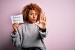 Young African American afro woman with curly hair holding paper with fake news message with open hand doing stop sign with serious and confident expression, defense gesture