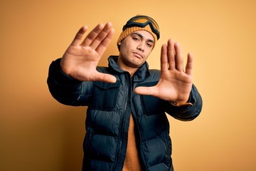 Wall Mural - Young brazilian skier man wearing snow sportswear and ski goggles over yellow background doing frame using hands palms and fingers, camera perspective